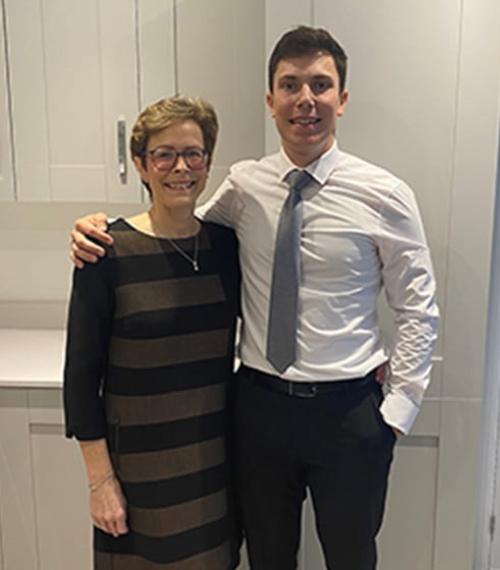 Mother in a black dress stood next to her son in a white shirt, blue tie and black trousers. He has his arm around her.