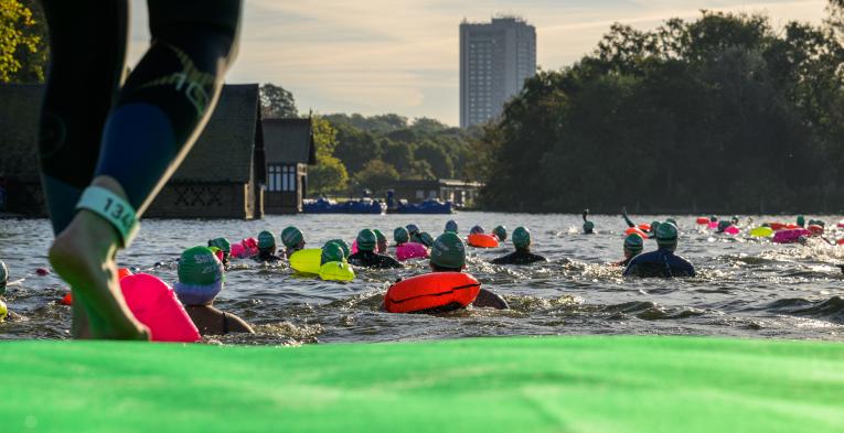 View of a swimmers legs as they walk into the serpentine. There are a group of swimmers in front of them already in the water with colourful floats swimming away from the bank. 