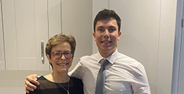 Mother in a black dress stood next to her son in a white shirt, blue tie and black trousers. He has his arm around her. 
