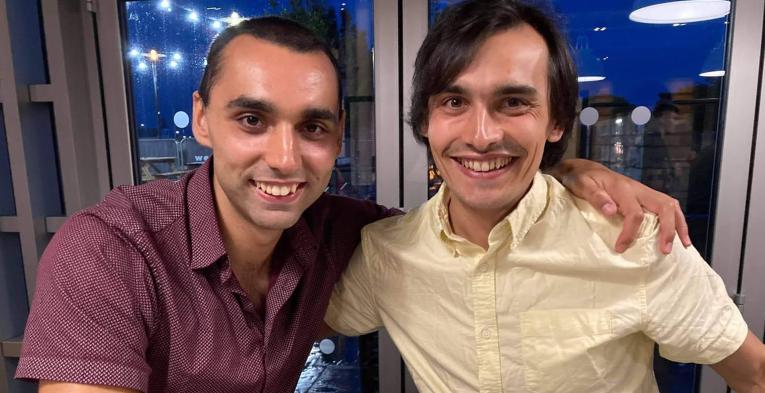 Two brother sat next to each other with their arm on each others shoulders 