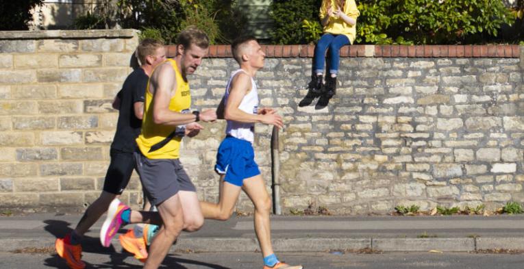 Men running on the road whilst a girl watches from the side, sitting on a wall.