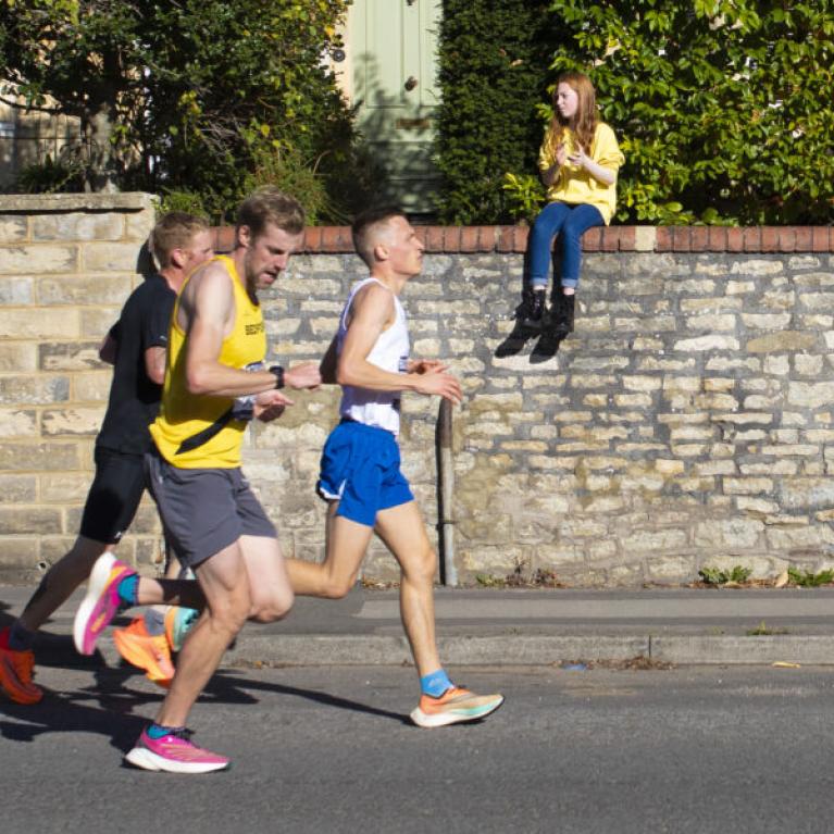 Men running on the road whilst a girl watches from the side, sitting on a wall.