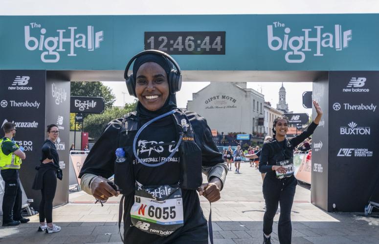 Smiling woman wearing a rucksack and headphones crosses the finish line. woman running behind her waves to the spectators.