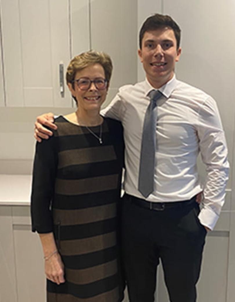 Mother in a black dress stood next to her son in a white shirt, blue tie and black trousers. He has his arm around her. 