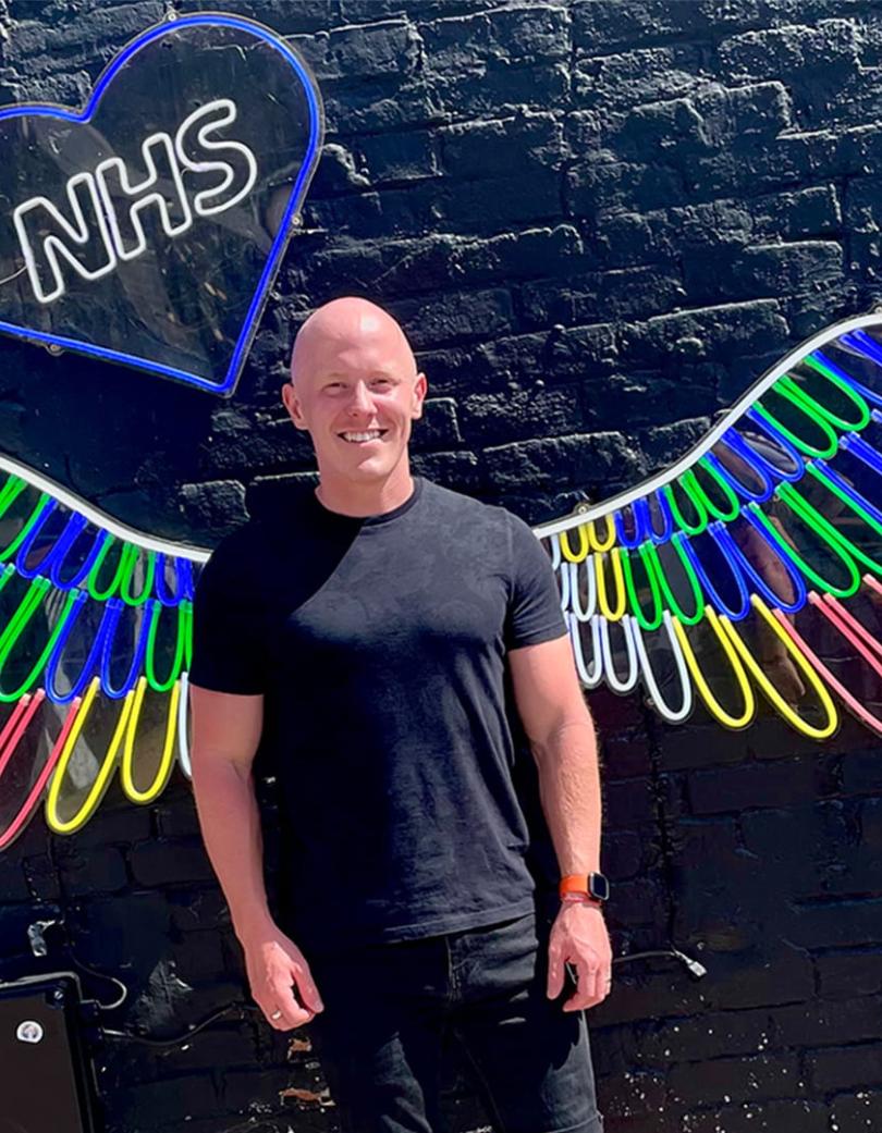 Person stood in front of a black wall with neon wings and NHS in a blue heart.